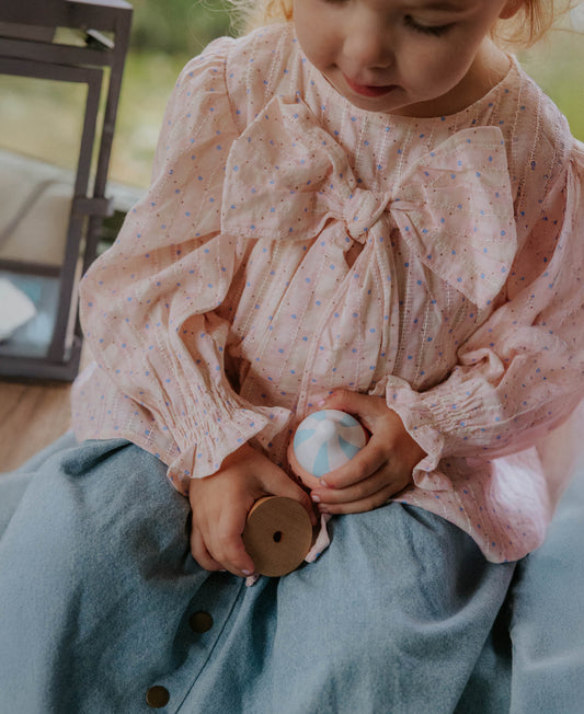 Child wearing the chalk pink bow blouse with pale blue trousers, holding a wooden toy