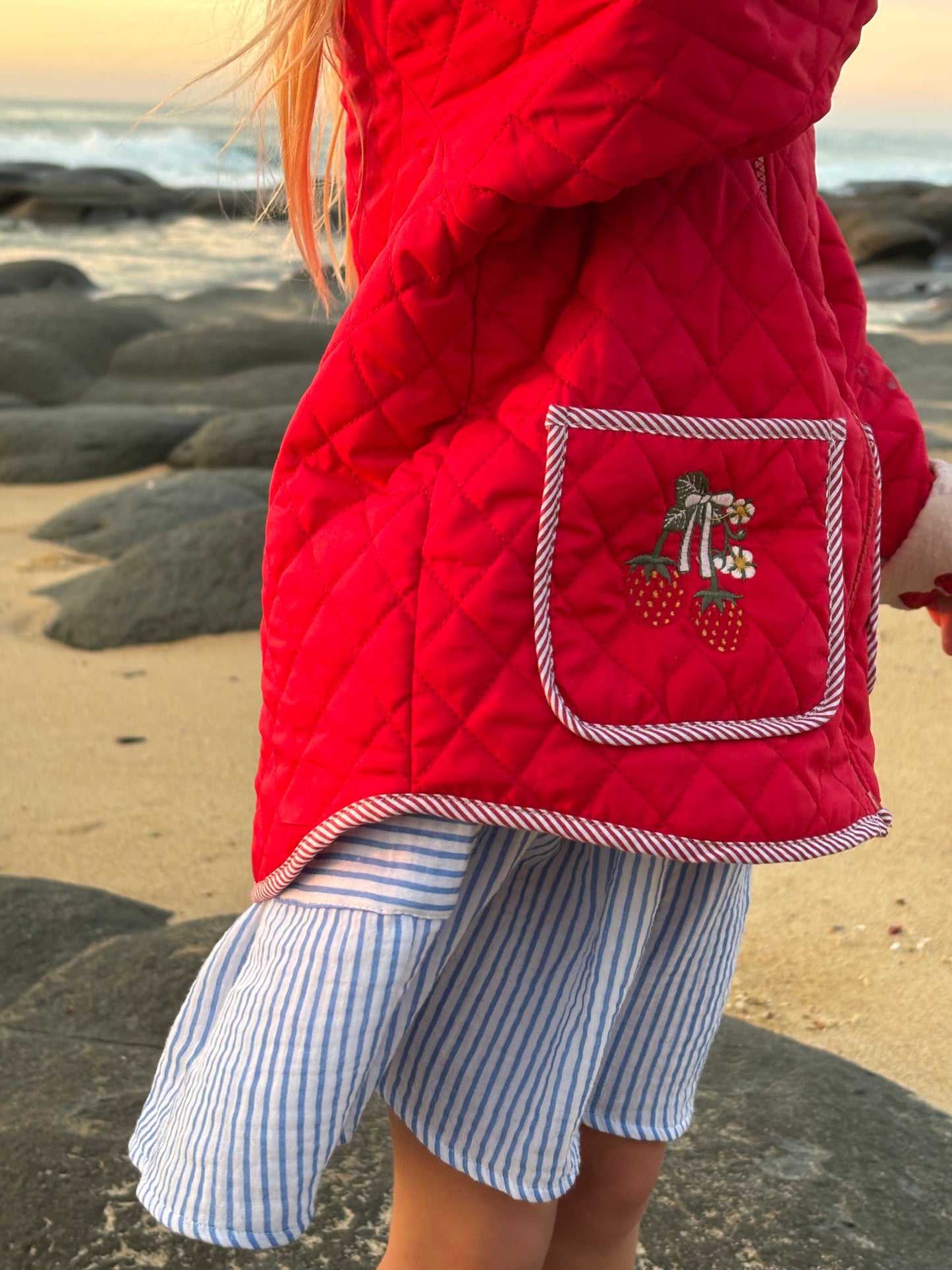 Close-up of the embroidered strawberry pocket on the red Kota jacket, worn at the beach