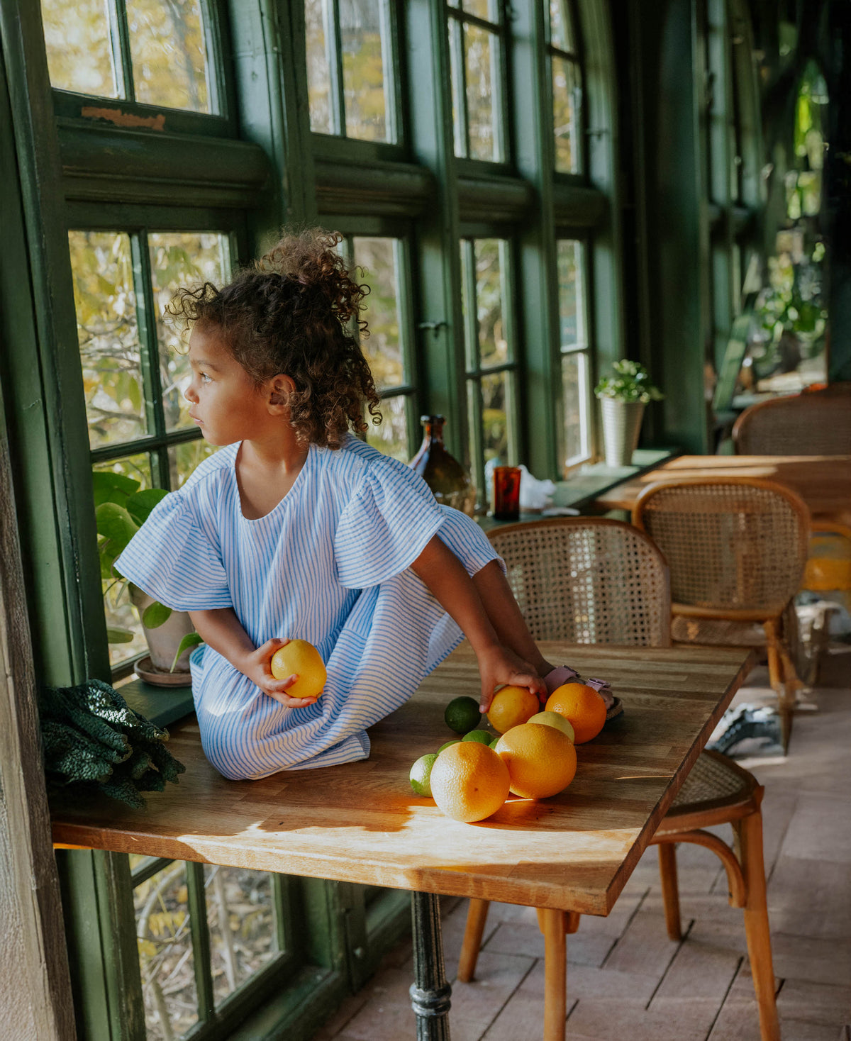 Child sitting on a table among oranges in the Cha Cha Dress in Marina Striped