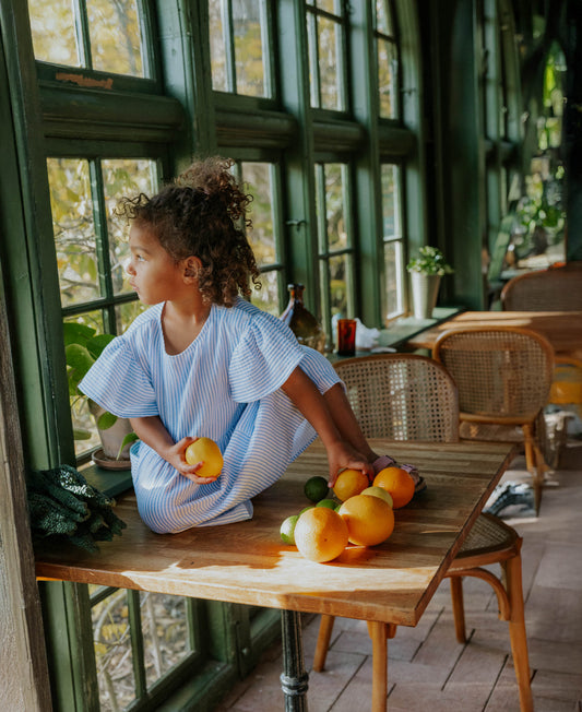 Child sitting on a table among oranges in the Cha Cha Dress in Marina Striped