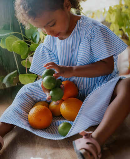 Child holding oranges in the lap of the blue striped Cha Cha Dress