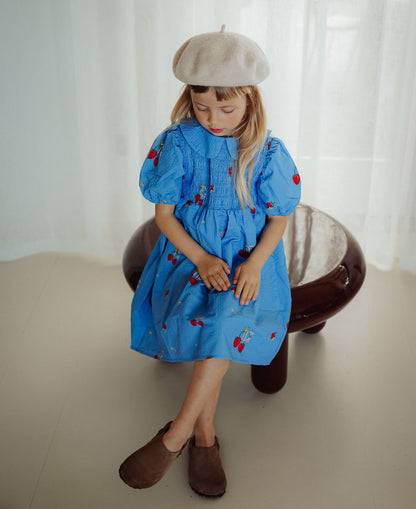 Child in a beret sitting on a stool in the blue strawberry-print Elma Dress