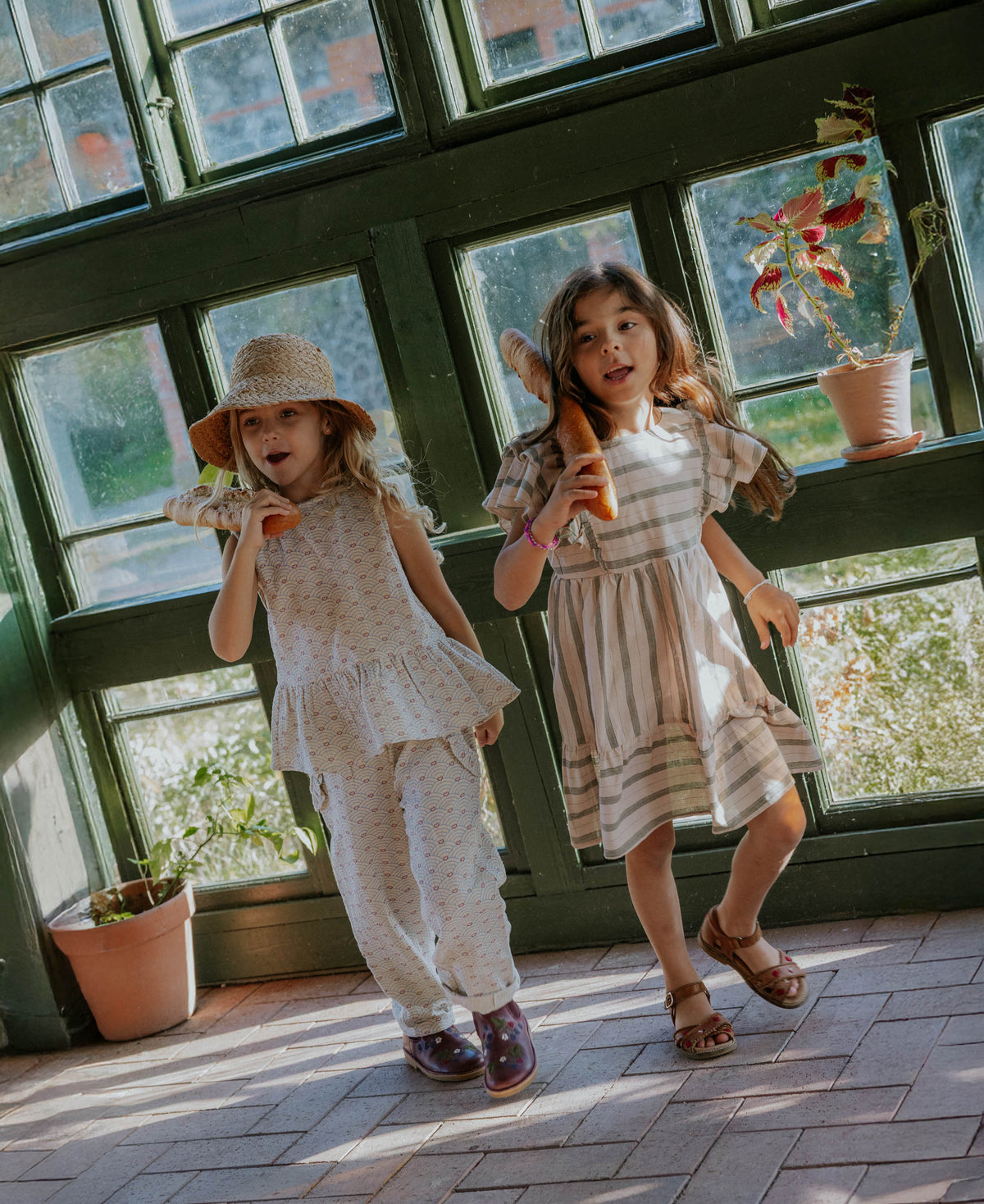 Two children in dresses standing in a sunlit room with large windows.