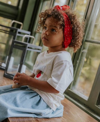 Child with a red headscarf sitting by a window in the Sommer Strawberry T-shirt