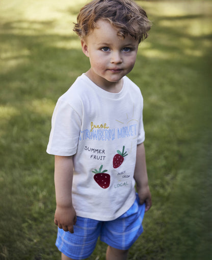 Child standing on grass in the white Sommer Strawberry T-shirt and blue checked shorts