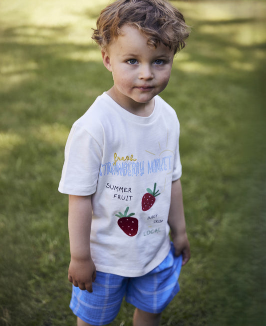 Child standing on grass in the white Sommer Strawberry T-shirt and blue checked shorts
