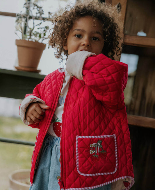 Child wearing the red quilted strawberry jacket open over a white top and denim skirt, standing outdoors