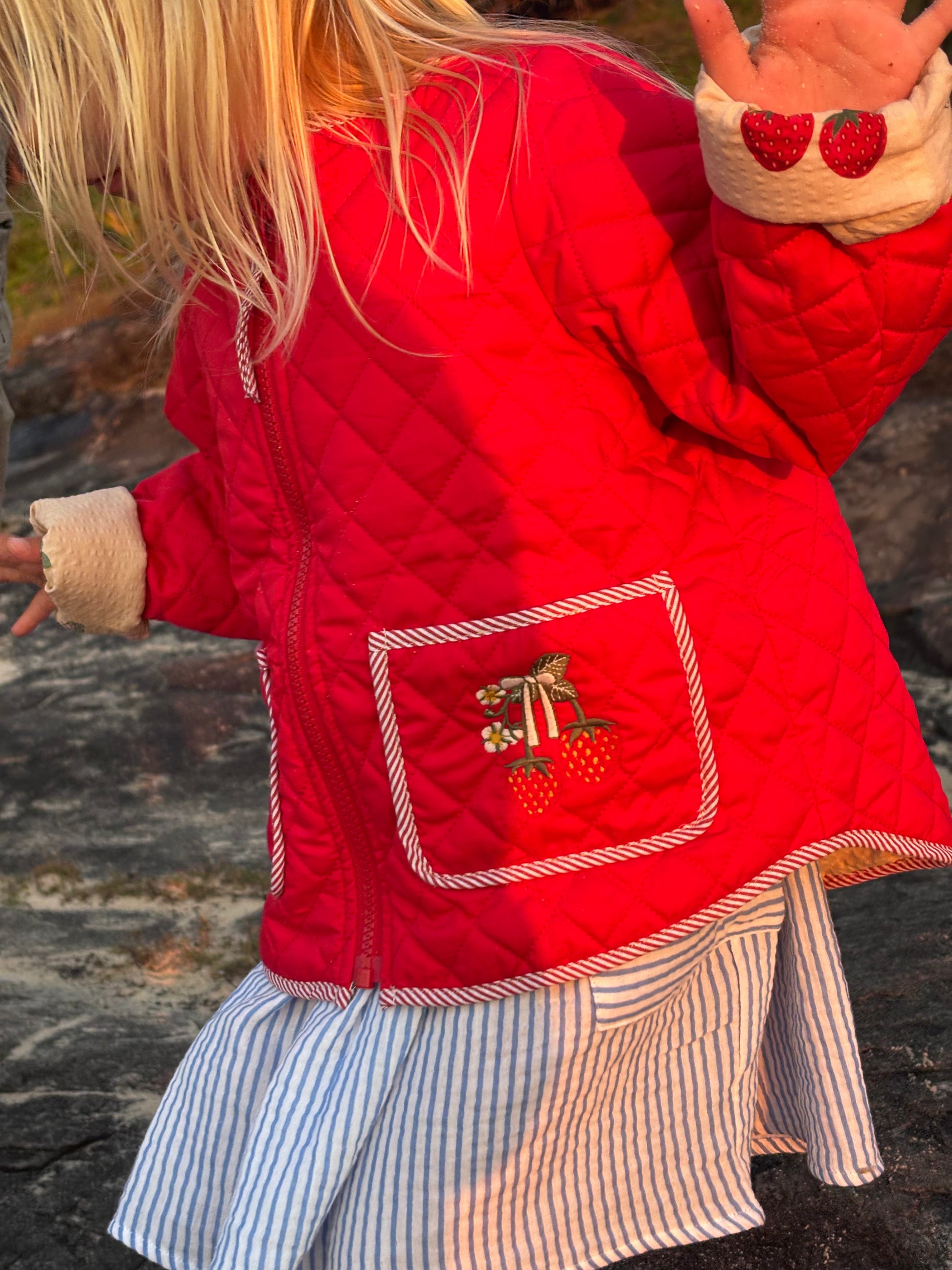 Child outdoors in the red quilted strawberry jacket over a blue and white striped skirt, seen from behind