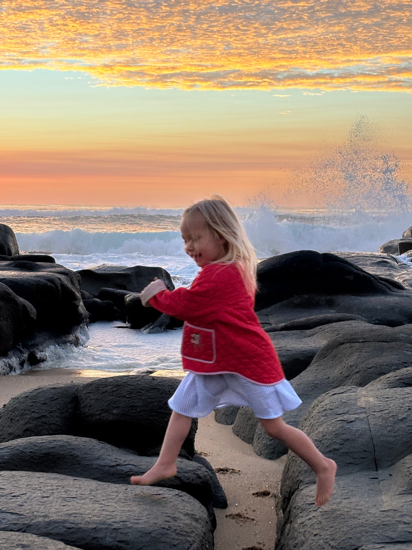 Girl running barefoot over beach rocks at sunset, wearing the red quilted jacket and a striped skirt