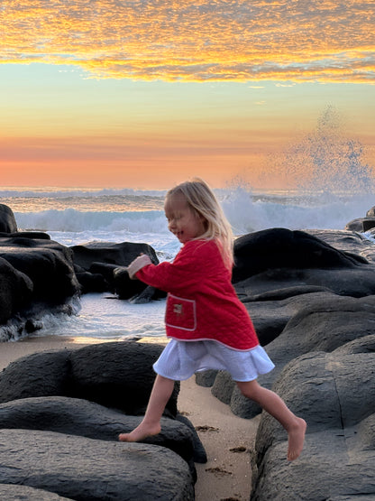 Girl running barefoot over beach rocks at sunset, wearing the red quilted jacket and a striped skirt