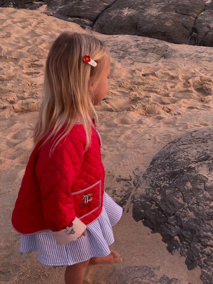 Girl walking on sand in the red quilted jacket and a blue and white striped skirt, side view