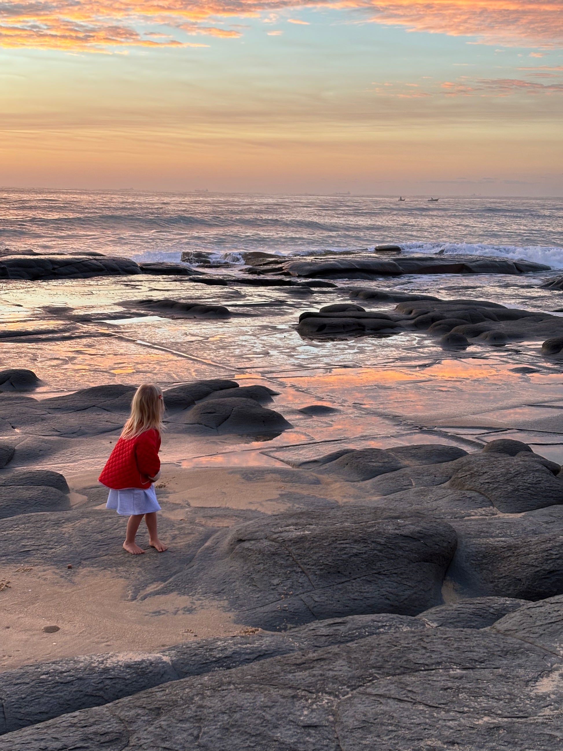 Child standing on a rocky beach at sunset in the red quilted jacket, looking out to the ocean