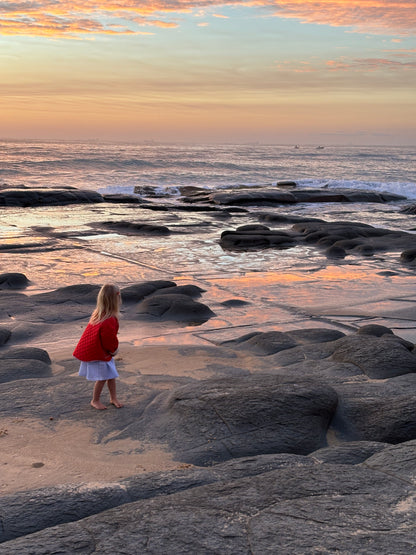 Child standing on a rocky beach at sunset in the red quilted jacket, looking out to the ocean